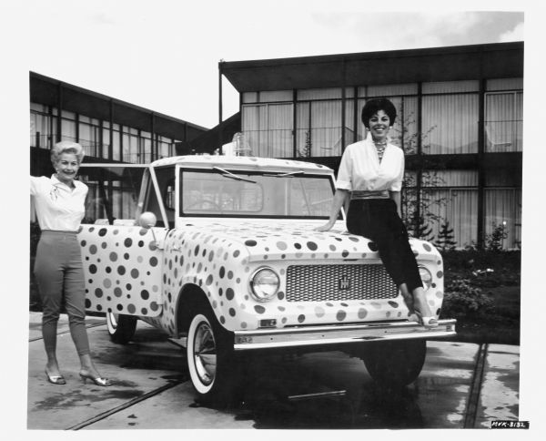Two women pose with an International Scout decorated in polka dots. The Scout is parked outdoors near a pool at what appears to be a motel or hotel.