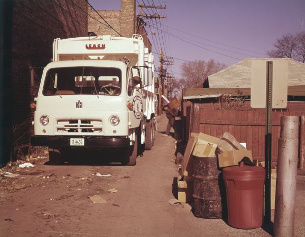Color photo of a man collecting garbage cans from an alleyway and emptying them into a truck with an IH logo on the front of the chassis and a Leach logo on the garbage truck.