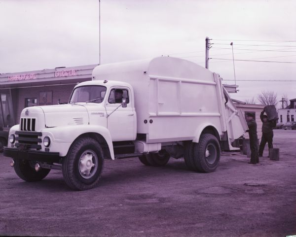Color photo of men unloading refuse into the back of an International garbage collection truck parked beside a restaurant. The neon signs on the restaurant building advertise hamburgers and chicken.