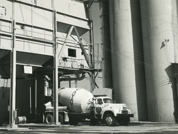 An International model RF-192 truck outfitted with a concrete mixer parked in front of industrial buildings. A man stands on the running board next to the passenger-side door of the truck.