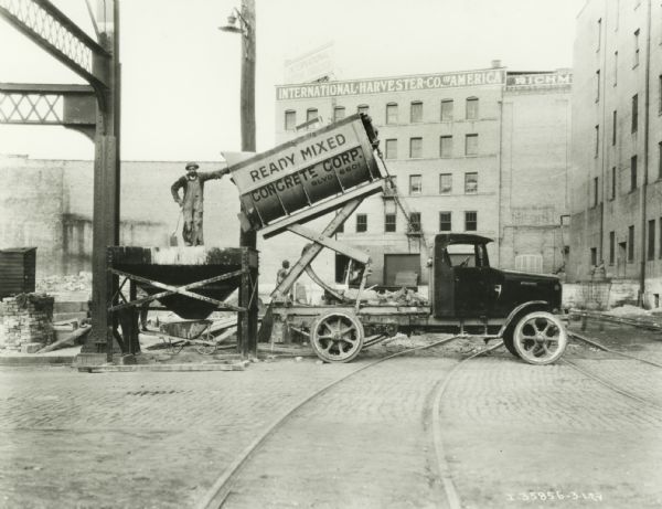 A man stands on a piece of equipment holding a shovel to move the contents of an International truck owned by the Ready-Mixed Concrete Corp. into a wheelbarrow standing below it. Buildings labeled: "International Harvester Co. Of America" are in the background, and another man is working in the yard.