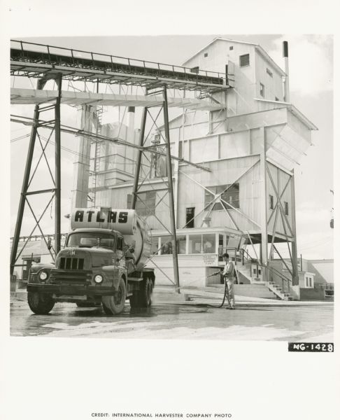 An International truck owned by Atlas Concrete Products, Inc. parked in front of industrial buildings. A man is standing on the right hosing off the truck. In the background another man is in a glassed-in office at the base of a tall industrial tower.