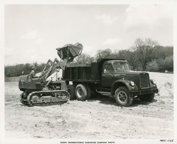 A man uses an International TD-6 crawler tractor (TracTracTor) with a Drott skid-shovel to load dirt into the bed of an International VF-190 dump truck.