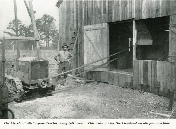 A man uses the power take-off on the Cleveland All-Purpose crawler tractor. A belt is running off of the tractor into a barn. The caption reads: "The Cleveland All-Purpose Tractor doing belt work. This work makes the Cleveland an all-year machine."