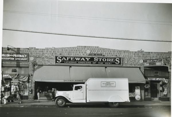 View across street of an International truck parked outside of a Safeway grocery store. The sign on the truck reads: "Meat Refrigerator Service; Safeway Markets." On the left side of the grocery store is a pharmacy, and on the right is Ada Marie Dry Goods and Beauty Shoppe.