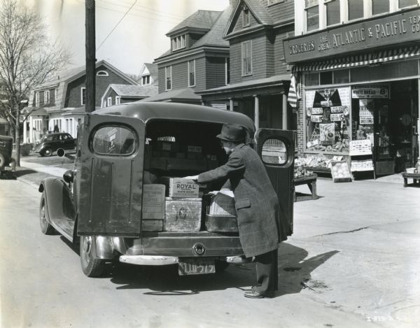 A man is loading boxes into the rear of an International C-1 panel truck owned by Standard Brands, Inc. The truck is parked alongside a street composed of residential buildings and The Great Atlantic & Pacific Tea Co.