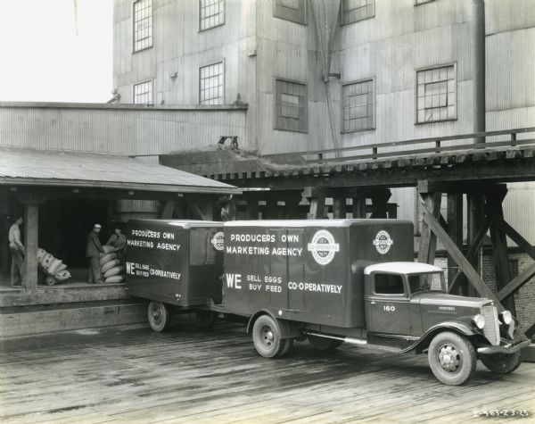 Men load bags from a loading dock onto the back of an International C-40 truck equipped with a trailer.