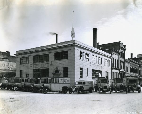 A fleet of trucks owned by the Tri-State Ice Cream Company lined up in front of the company building. A sign in the shape of a milk bottle stands on top of the building. On the right is a sign on a building for the La Crosse Steam Laundry.