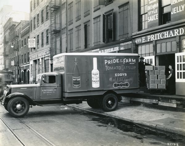 An International truck owned by E. Pritchard, Inc. makes a delivery to a loading dock at the company warehouse on a city street. The truck advertises Pride of the Farm tomato catsup and Eddys Old English Style Sauce.