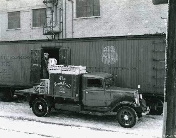A man loads fruit crates onto the back of an International truck owned by the Scott Fruit Company Limited. Behind him is a ventilated refrigerator railroad car on railroad tracks next to a factory building.