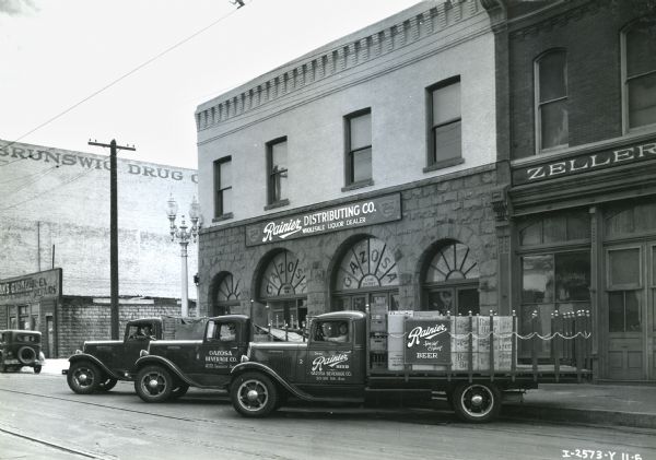 Three of the five International trucks, owned by the Gazosa Beverage Company, are parked at an angle outside of the Rainier Distributing Company. Men wearing hats sit in the driver's seat of each truck looking at the camera. The side of the truck has a "Rainier" beer sign.