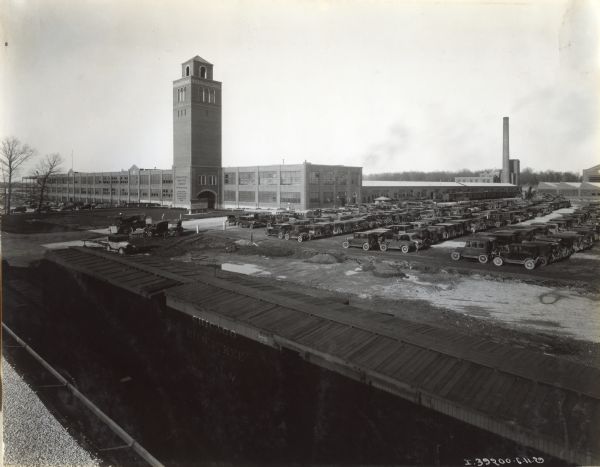 Elevated exterior view of International Harvester's Ft. Wayne Works, including the factory's tower and a parking lot full of cars. Railroad boxcars are in the foreground.