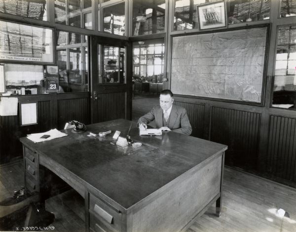 A man, probably a factory manager, sitting at a large desk in an office at International Harvester's Ft. Wayne Works. A large map of the United States is hanging on the wall behind him with pins in it. A framed photograph of the "First Truck Produced at Ft. Wayne Works" is hanging on another wall. Other offices are visible through windows in the background.