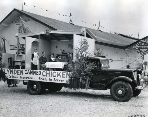 Men and women pose in a household scene constructed on an International C-line truck for use in a parade on Egg Day at the Western Washington Fair. The parade float was constructed by the Washington Cooperative Egg and Poultry Association and the sign on the side of the truck reads: "Lynden Canned Chicken. Delicious. Economical. Ready to Serve."