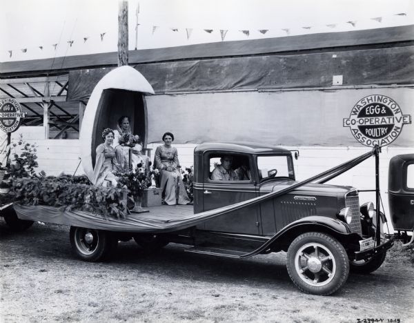 Women ride on a parade float on the bed of an International C-line truck during Egg Day at the Western Washington Fair. Two men are riding inside the cab of the truck.