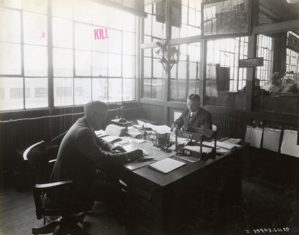 Two men, possibly clerks, sit on either side of a desk working on papers in an office at International Harvester's Ft. Wayne Works. There is a phone, stamps, in-out box and other office equipment. Windows to the outside are behind them; another office is visible through windows along the wall on the right.