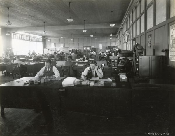 A large office full of male and female clerks at desks, working on papers and talking on the phone in the Time Keeping Department at International Harvester's Ft. Wayne Works.