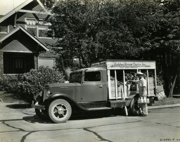 A man unloads milk bottles from the back of an International C-30 truck owned by Golden Arrow Dairy, Inc. A house is in the background.