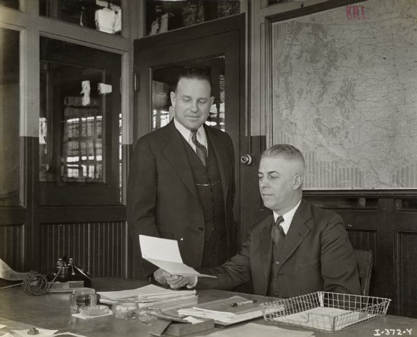 Assistant Superintendent C.M. Harrison (left) and Superintendent E.C. Lutz look at a document in an office at International Harvester's Ft. Wayne Works. A large map of the United States with pins in it is hanging in the background.