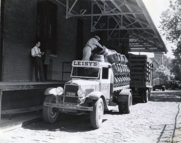 A man loading barrels onto the body of an International truck owned by Leisy's Brewery as another man is looking on from a loading dock.