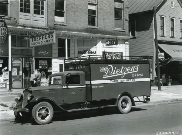 An International C-35 truck owned by Dietzen's Bakery parked on a city street alongside a curb in front of Toepfer's Groceries and Meats.