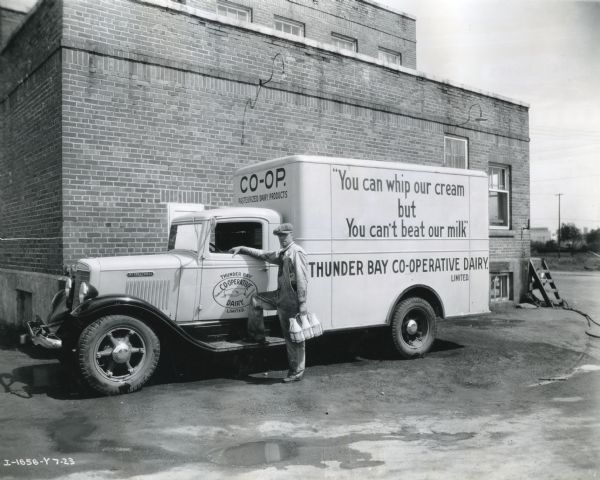 A delivery driver holds a container of milk bottles while standing next to an International C-30 truck owned by Thunder Bay Co-Operative Dairy Limited. The photograph was taken in Port Arthur, Ontario, Canada and the text on the truck reads, "You can whip our cream but You can't beat our milk."