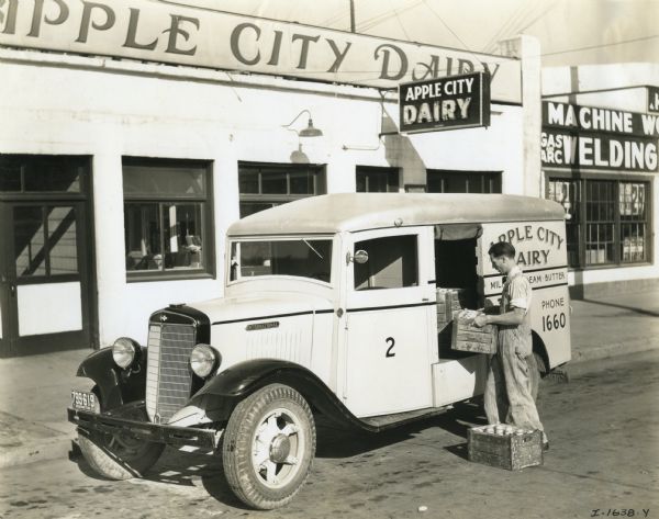A man loads wooden crates full of milk bottles into the back of an International C-20 truck owned by Apple City Dairy. The dairy building is in the background.