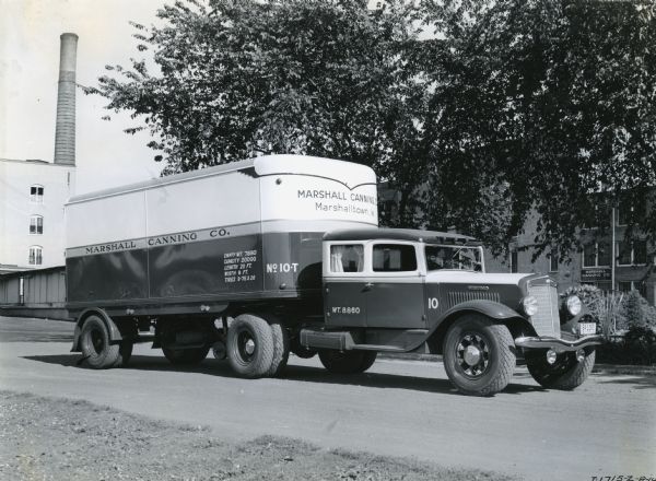 Three-quarter side view of an International Model C-55 truck with a sleeper cab and Fruehauf semi-trailer owned by the Marshall Canning Company. The factory building is in the background.