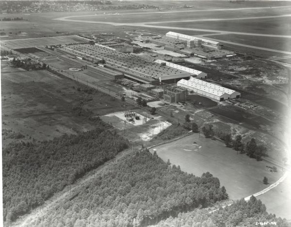 Aerial view of factory buildings and grounds at International Harvester's Louisville Works. Roads, fields and trees surround the factory. In the foreground there appears to be a golf course. In the far distance is a town.