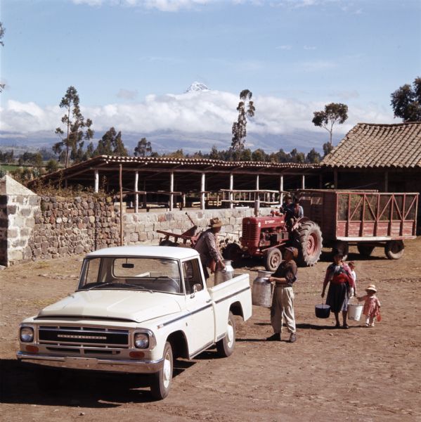 Slightly elevated view of men loading milk pails onto the bed of an International pickup truck, while a woman carrying buckets is walking by, accompanied by two children. In the background a man on a tractor is pulling a wagon.