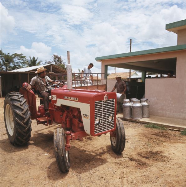 Color photo of a man siting on a McCormick 434 tractor as two men load milk pails onto a wagon. They are standing next to a building.