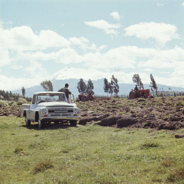 Color photo of a man standing beside an International pickup truck while watching two tractors work in a field. A mountain range is in the background.