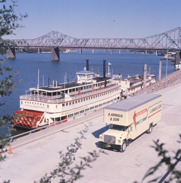 Color photo of an International Harvester model 1700 truck owned by A. Arnold and Son Transfer and Storage Co. by the Ohio River. On the river is a sternwheel steamboat named "Belle of Louisville." Two bridges are in the background. The photo caption reads "Eye-pleasing merger at Louisville of one of Arnold’s 1700's, a sternwheel steamboat, Ohio River and bridges provided this issue's cover."
