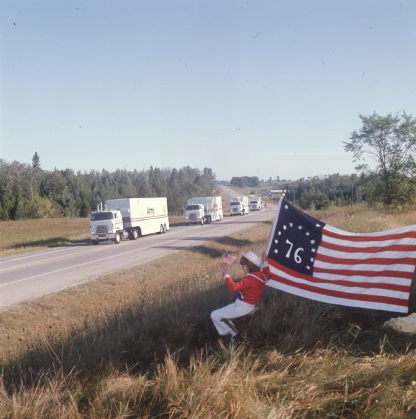 Seven-year-old LuRae Criscione watches the International Harvester United States Armed Forces Bicentennial Caravan. LuRae is dressed in red, white, and blue clothes with a large '76 flag in one hand and a miniature flag in the other. She is sitting by the side of a road while watching the procession of International Transtar II heavy-duty trucks. The trucks were part of an eighteen month caravan to honor the United States Bicentennial and armed forces. The caption for the color photo reads: "Since caravan made debut in July before Washington, the above scene has been common sight, coast to coast."