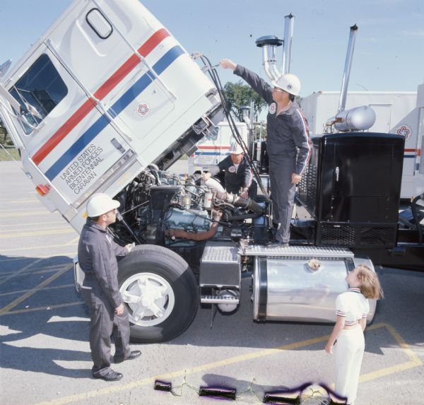 Color photo of three men and a small girl named LaRue Criscione examining a truck engine. The truck is an International Transtar II, and part of the International Harvester United States Armed Forces Bicentennial Caravan. The men are in coveralls and hardhats, while LaRue is dressed in a white outfit with red and blue bands. The photo caption reads: "On-site examinations of newly developed IH V-800 diesel engines, standard power for 16 display rigs, are daily ritual. Here, crew manning Caravan Two's Air Force vehicle, fascinated viewer LuRae Criscione, 7, of Berwyn, Illinois, as servicemen tilted cab of Transtar II shortly after in-transit procession was photographed from the air by Air Force T/Sgt. Deal Toney."