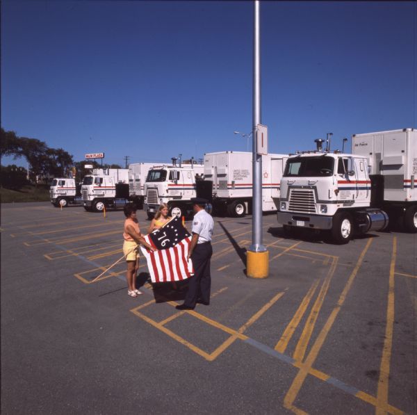 View of a woman, young woman, and a man in a military uniform are standing with a '76 flag. In the background are four International Transtar II heavy-duty trucks. The trucks were part of an eighteen month caravan to honor the United States Bicentennial and armed forces. A sign in the background reads, "M&M Plaza." The photo caption reads, "As evident as Menominee, Mich., engagement, flags of Revolution are frequently displayed by people welcoming the caravan."
