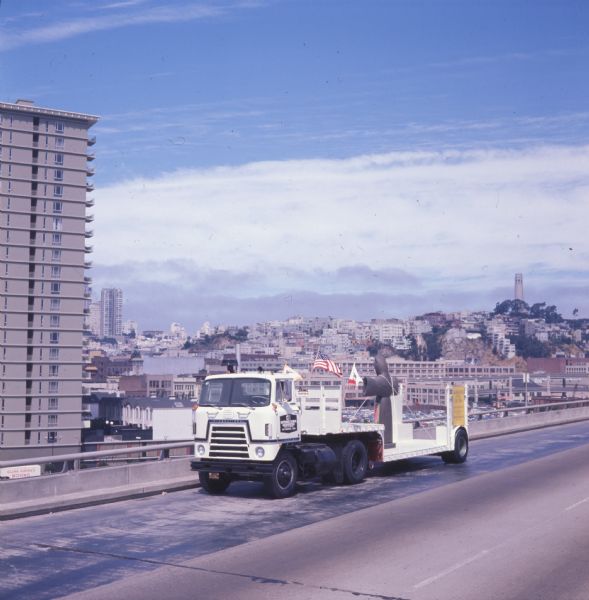 Color photo of an International C-O 4070A Transtar truck hauling the metal statue "St. Francis of the Guns" on a trailer down a San Francisco highway. The statue represents St. Francis of Assisi and was created by San Francisco sculptor Beniamino Bufano. The photo caption reads: "'Standing' in lowboy, St. Francis of the Guns seemed to be blessing city of San Francisco as rig started down the freeway."