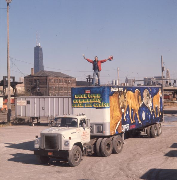 View of a man in a red jacket holding paint brushes and a palette while standing atop a truck trailer. The side of the trailer is painted with a mural of four endangered large cat species with "Save our cats" written above it. The animals depicted are possibly a lion, tiger, lynx, and puma. The front of the trailer reads, "West Town Community Youth Art Center" in large yellow and green letters. The man on top of the trailer is Ricardo Alonzo, muralist and director of Chicago's West Town Community Youth Art Center. The trailer is pulled by a white International Harvester F-2010A truck. The door of the truck reads, "Operated by March Tranport Co., 3401 W. Pershing Rd., Chicago Ill." In the background is the Willis Tower (formerly named and still commonly referred to as Sears Tower).