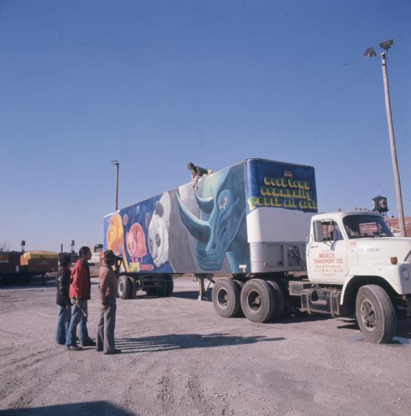 Three men are looking at a trailer with a woman posing on roof holding a paintbrush to a painted mural. One of the men is Ricardo Alonzo, muralist and director of Chicago's West Town Community Youth Art Center. The woman is Ruth Felton. The front of the trailer reads: "West Town Community Youth Art Center" in large yellow and green letters. The side of the trailer is part of a series of murals depicting endangered land animals, and shows a rhinoceros with a tear in its eye, a panda, and two other animals. Attached to the front of the trailer is a white International Harvester F-2010A truck. The door of the truck reads: "Operated by March Tranport Co., 3401 W. Pershing Rd., Chicago Ill."