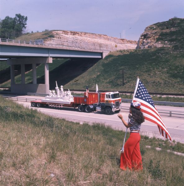 View looking down hill towards a girl holding an American flag near a highway. Jane Leeney, in red, white, and blue pants, shirt, and hat is watching a truck pulling a trailer with a limestone sculpture of General George Washington crossing the Delaware River. The truck is a red, white, and blue International Harvester COF-4070B truck. The truck has just gone through an underpass, which is in the background.