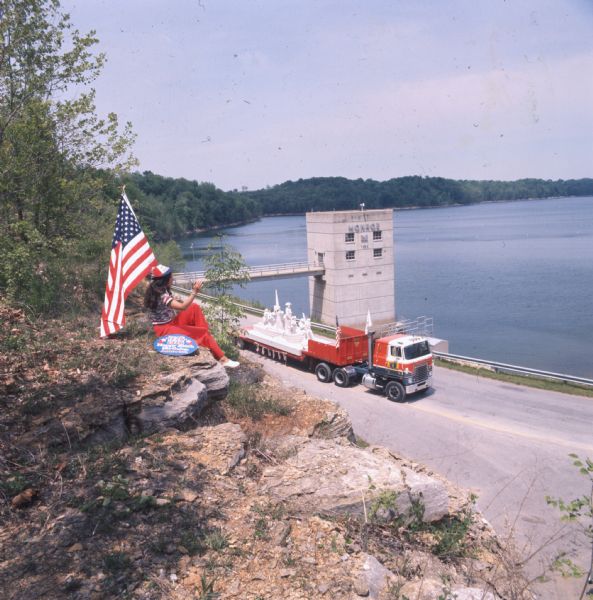 Jane Leeney, in red, white, and blue pants, shirt, and a hat is sitting on a cliff. She is holding a large flag, and a sign beside her reads, "1776-1976 Happy 200th Birthday America." She is watching a truck pulling a trailer with a limestone sculpture of General George Washington crossing the Delaware River. The truck is a red, white, and blue International Harvester COF-4070B truck. The truck is driving on a road beside a body of water, probably a lake, with a large square concrete building in it connected by a walkway. The structure reads "Monroe," and may refer to Monroe, Indiana.