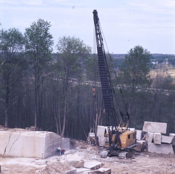 Two workers and a large yellow crane at a limestone quarry. There are large blocks of roughly cut limestone, and numerous trees are in the background. The quarry is likely the David Elliott Stone Company of Bedford, Indiana.