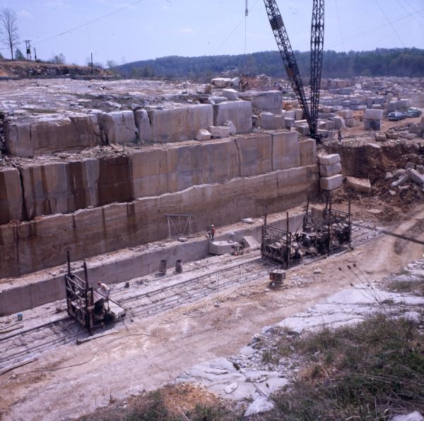 Color photo of workers cutting blocks of limestone in a quarry. There are what appear to be moveable platforms on tracks at the bottom of the face of the area being cut into, and workers standing on these platforms. Workers can also be seen standing on a large block of stone at the bottom of the face. A large crane-like structure, and numerous rough cut blocks of stone, are in the background. The quarry is the David Elliott Stone Company's Shawnee deposit.