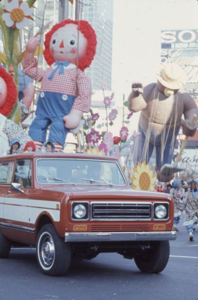 Thanksgiving Day Parade, either Macy's Parade in Manhattan, New York, or Gimbels' Parade in Philadelphia, Pennsylvania. A red and white International Harvester Scout 4x4 is towing a parade float with Raggedy Ann (partially out of frame) and Andy in a field of flowers. Children in costume are on the front of the float. Other members of the parade are walking beside the float, and in the background is a second float with a large Smokey the Bear character with his customary hat and shovel.