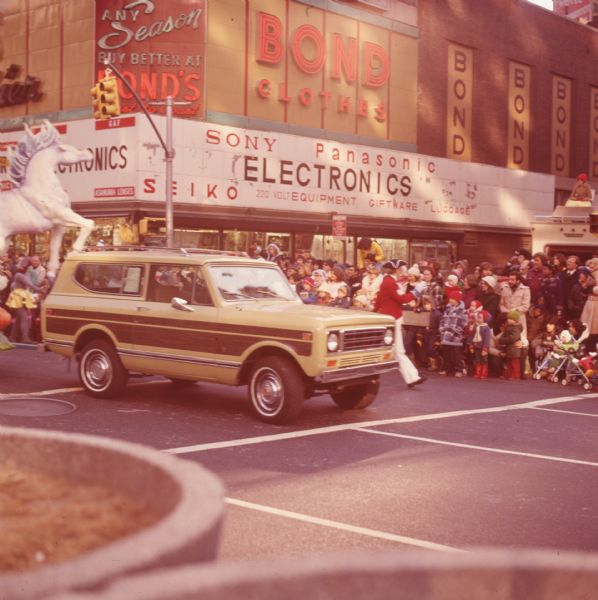 Thanksgiving Day Parade, either Macy's Parade in Manhattan, New York, or Gimbels' Parade in Philadelphia, Pennsylvania. A tan and brown International Harvester Scout 4x4 is pulling a float (possibly a Cinderella-themed float also seen in another image). The float cannot be seen entirely, but a large white horse on the front is in frame. A man in white pants, red jacket, dark blue tri-corner hat, and white gloves with blue trim is walking beside the Scout, and waving to spectators. Spectators can be seen under large signs reading "Bond Clothes," "Any Season Buy Better at Bond's Coast to Coast," Bond," and "Electronics, Sony, Panasonic, Seiko, 220 Volt, Equipment, Giftware, Luggage."