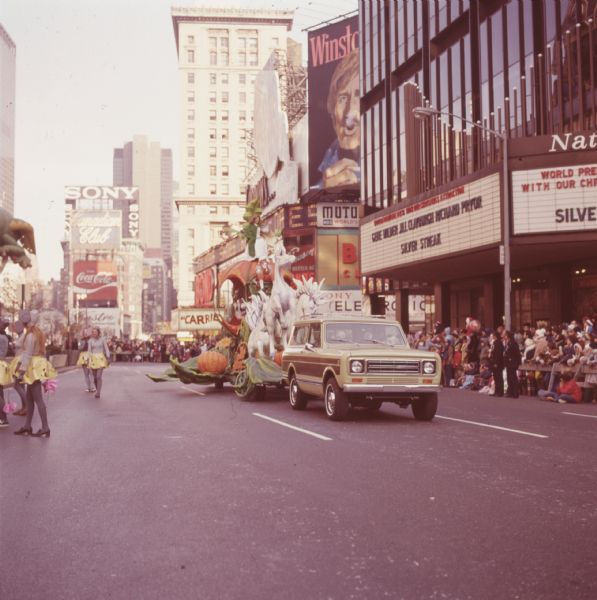 Thanksgiving Day Parade, either Macy's Parade in Manhattan, New York, or Gimbels' Parade in Philadelphia, Pennsylvania. A tan and brown International Harvester Scout 4x4 is pulling a float with a Cinderella theme. The float has several white horses pulling a pumpkin carriage with driver in orange and gray livery, and a woman in a white gown and green shawl sitting on top. A sign on the side appears to read "Dr. Pepper." Other signs in the background and along the street advertise Sony, Coca-Cola, Canadian Club, Winston cigarettes (with image of man smoking a cigarette), and the motion pictures Carrie and Silver Streak. Women in gray costumes with yellow skirts (possibly representing mice) walk along one side of the float, and spectators and uniformed police officers can be seen on the other side.