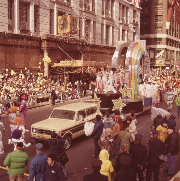 Macy's Thanksgiving Day Parade. The Macy's storefront can be seen in the background. A yellow and brown International Harvester Scout 4x4 is towing a float for 99x FM radio station. The float features a blue, green, yellow, red, and reflective arch with several similarly colored stars. Women in white dresses are dancing on the front of the float, along with a man in a white suit who has a microphone and stand. Men in white suits with white top hats are on the side of the float. People with either orange or purple colored hair and metallic suits are walking in front of the float, along with an African-American man in a red hat with clothing made of alternating blue and red squares with white dots. Spectators, uniformed police officers, and camera crews can all be seen around the float.