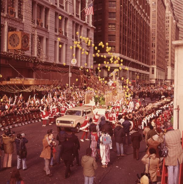 Macy's Thanksgiving Day Parade. The Macy's storefront can be seen in the background. A cream and white International Harvester Scout 4x4 is pulling a float with a Santa Claus theme. On the float, a group of reindeer are rising at an angle with Santa Claus in a sleigh behind. On the side of the float are children dressed in green as elves, and women in white clothes with red skirts and capes with white trim. Yellow balloons are rising into the air around the float. Alongside the float is a band in what appears to be Scottish dress, as well as spectators, camera crews, and uniformed police officers. Some of the band members are holding flags.