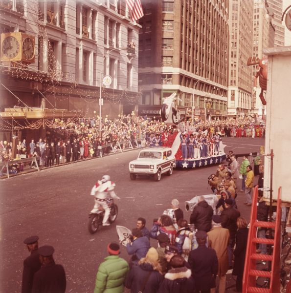 Macy's Thanksgiving Day Parade. The Macy's storefront can be seen in the background. A white and brown International Harvester Scout 4x4 is pulling a float with a motorcycle daredevil (probably Evel Knievel) theme. The float has a white motorcycle constructed to look like it has just jumped off a ramp, with adults and children on the side in jumpsuits of either red, white, or blue. The jumpsuits have a "V" shape down the chest and wide belts with stars. A person driving a motorcycle is facing towards the Scout and float. In the background a float towing a balloon depicting cartoon character Bullwinkle J. Moose can be seen. Spectators, uniformed police officers, and camera crews can all be seen around the float.