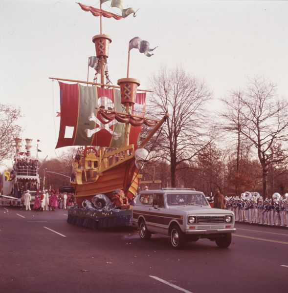 Color photo of a Thanksgiving Day Parade, either Macy's Parade in Manhattan, New York, or Gimbels' Parade in Philadelphia, Pennsylvania. A blue and brown International Harvester Scout 4x4 is pulling a float built to look like a pirate ship. The pirate ship has a large skull on the front, and a picture of a skull and crossbones on the sail. A red lobster-like creature with a yellow beard and what may be a blue bandana is rising out of the water at the base of the float. Behind the float is a group of men in white suits with white top hats and women in pink dresses with black trim and hats. Further back is a float with two levels, red, white, and blue striped pillars, and men in what may be Confederate soldier uniforms. A band can be seen on the side of the street, and trees are in the background.